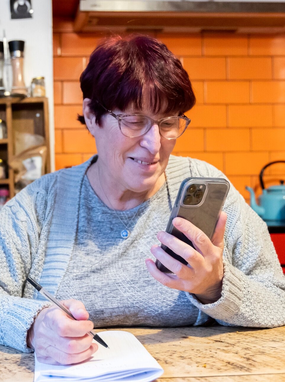 Woman looking at her smartphone while taking notes. Sat in kitchen.